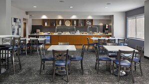 Zona pranzo moderna con tavoli e sedie, una cucina sullo sfondo e un'ampia finestra a destra.