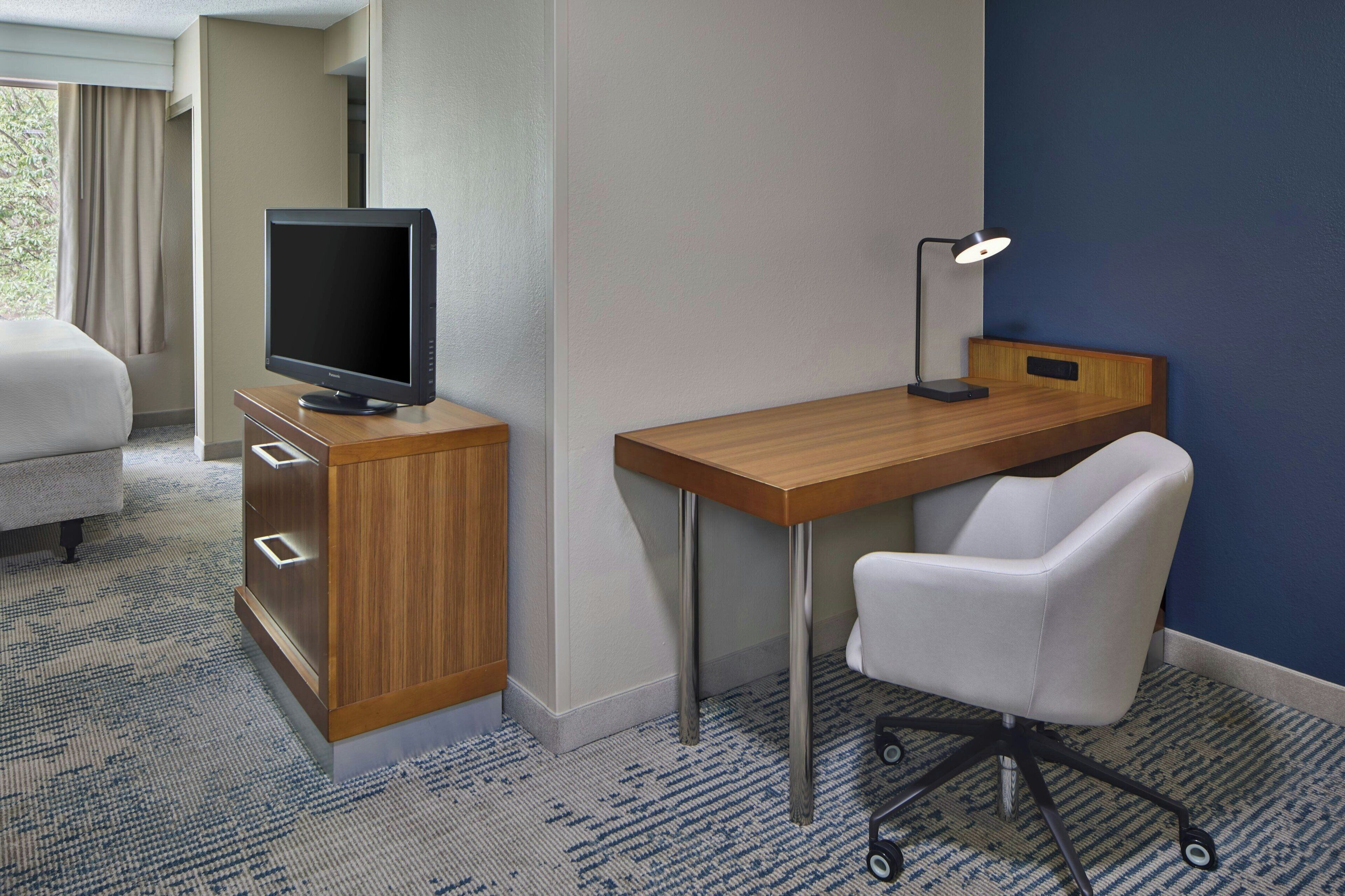 Een hotelkamer met een houten bureau, een witte bureaustoel, een televisie op een kast en een lamp.