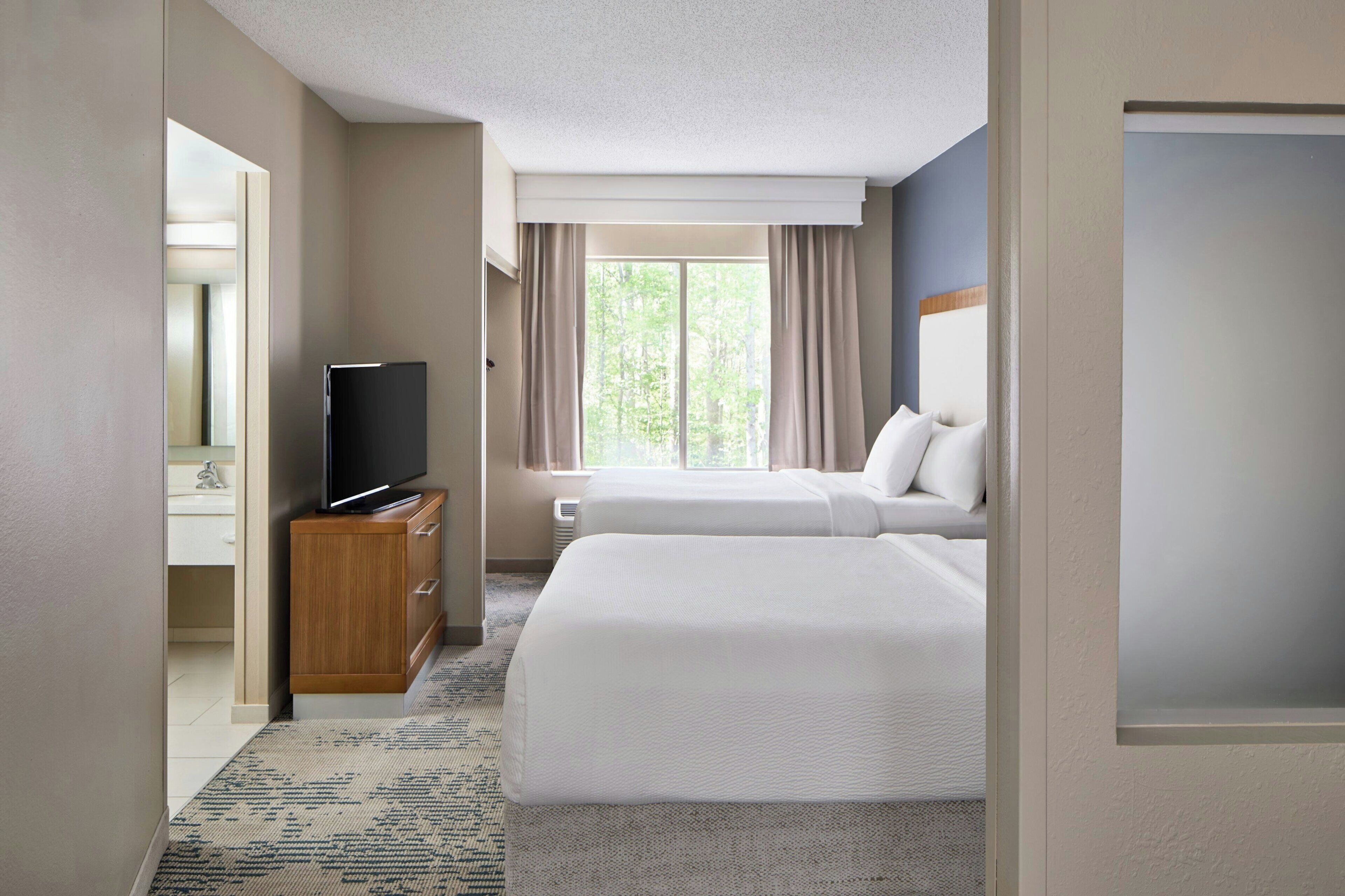Habitación de hotel con dos camas, un televisor sobre un mueble de madera, una ventana con cortinas y un baño visible a través de una puerta abierta.