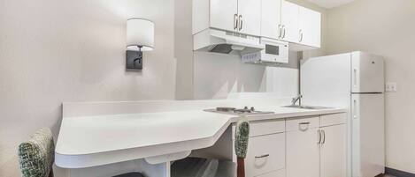 Cocina compacta con armarios blancos, microondas, refrigerador, lavamanos y un pequeño comedor con una silla y mesa.