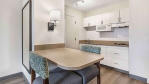 Una cocina compacta con armarios blancos, un microondas y una pequeña encimera. Hay una mesa de comedor con dos sillas, una de ellas con un cojín estampado.