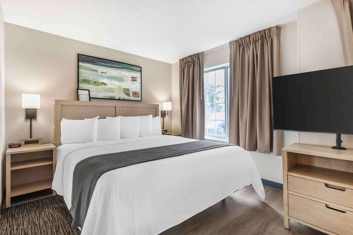 Habitación de hotel con una cama grande, dos mesitas de noche con lámparas, un televisor de pantalla plana sobre un mueble, un cuadro en la pared y una ventana con cortinas.