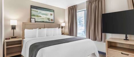 A hotel room with a large bed, two bedside tables with lamps, a flat-screen TV on a stand, a painting on the wall, and a window with curtains.