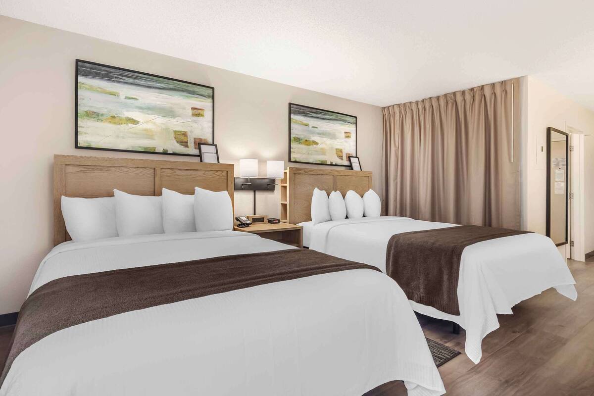 Habitación de hotel con dos camas, un cabecero de madera, una mesita de noche con lámpara y dos cuadros abstractos en la pared.