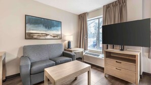 Una sala moderna con un sofá gris, una mesa de centro de madera, un televisor de pantalla plana, una mesita de noche con lámpara, y un ventanal con cortinas.