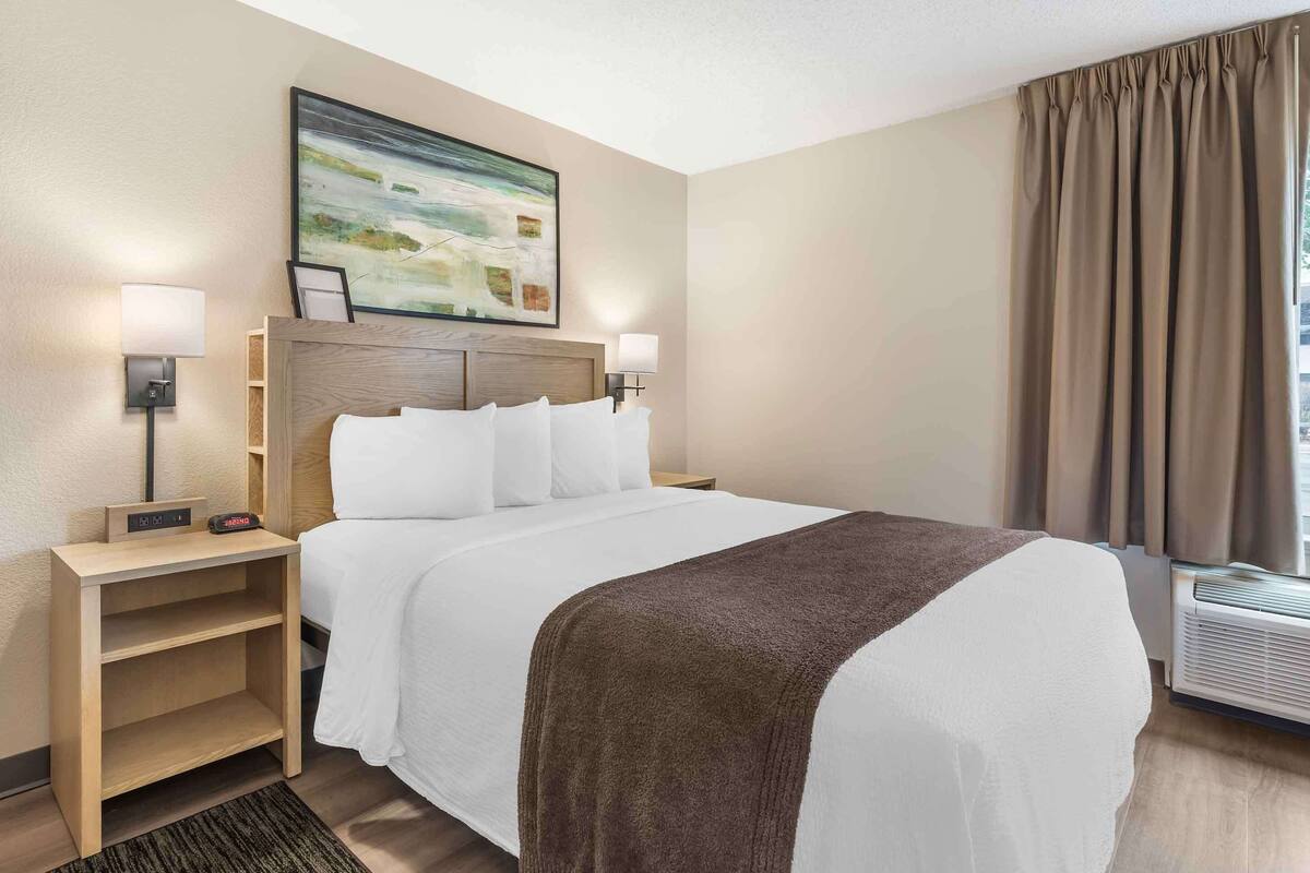 Habitación de hotel con cama, lámparas de noche, mesita de noche, un cuadro en la pared y una ventana con cortinas.