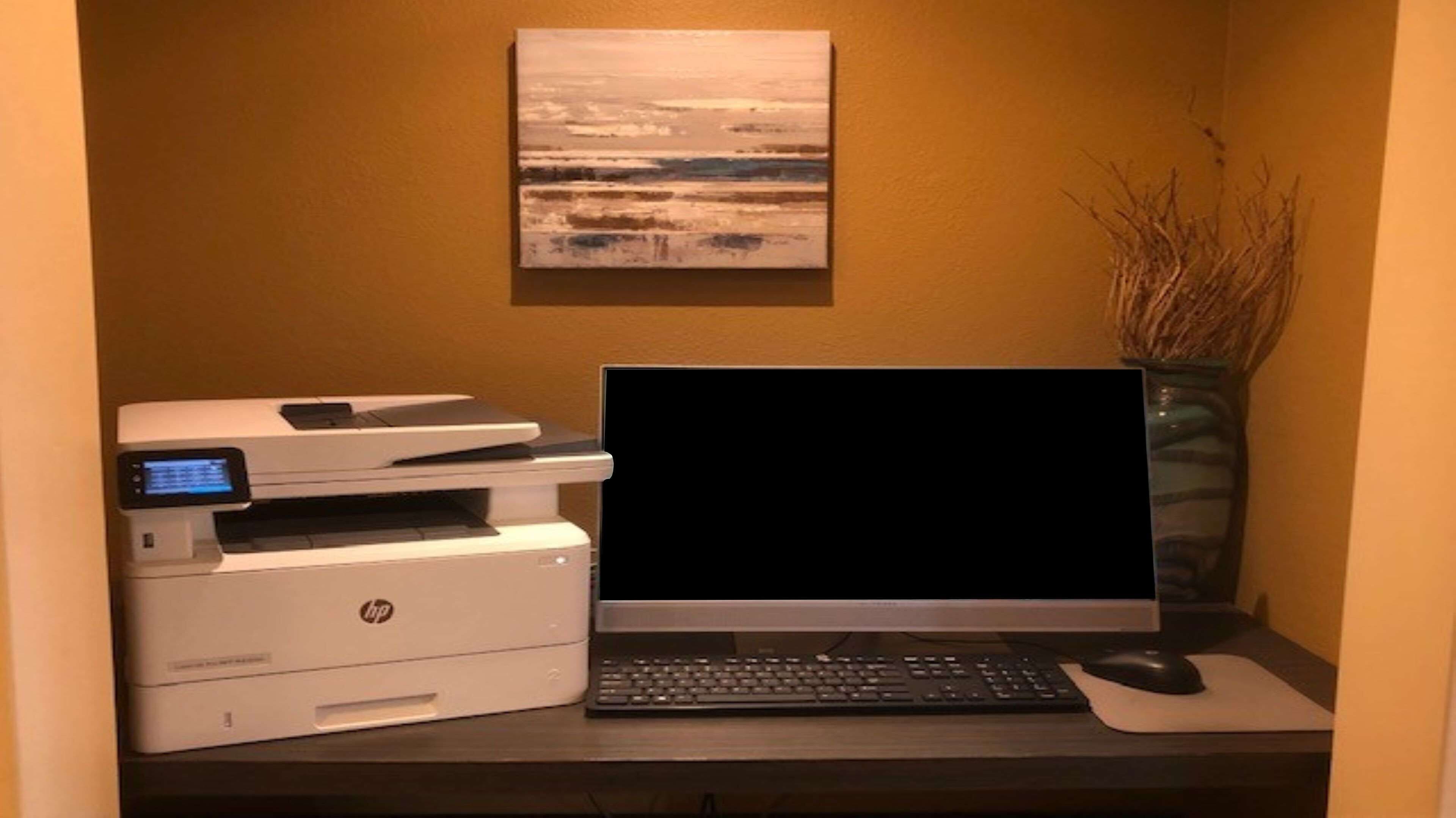 Un espacio de oficina en casa con una impresora, una computadora portátil, un teclado y un ratón sobre un escritorio, con un cuadro en la pared y un jarrón con ramas a la derecha.