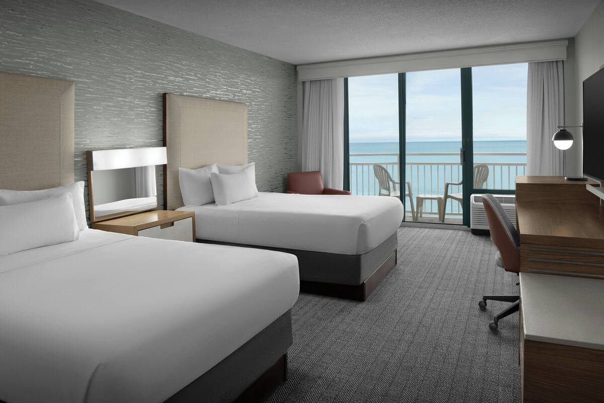 A hotel room with two beds, a desk, a chair, and a view of the ocean.