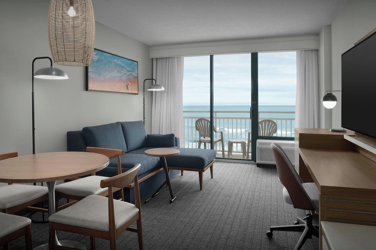 A hotel room with a sofa, a round table, a desk, and a TV. There is a large window with a view of the ocean.