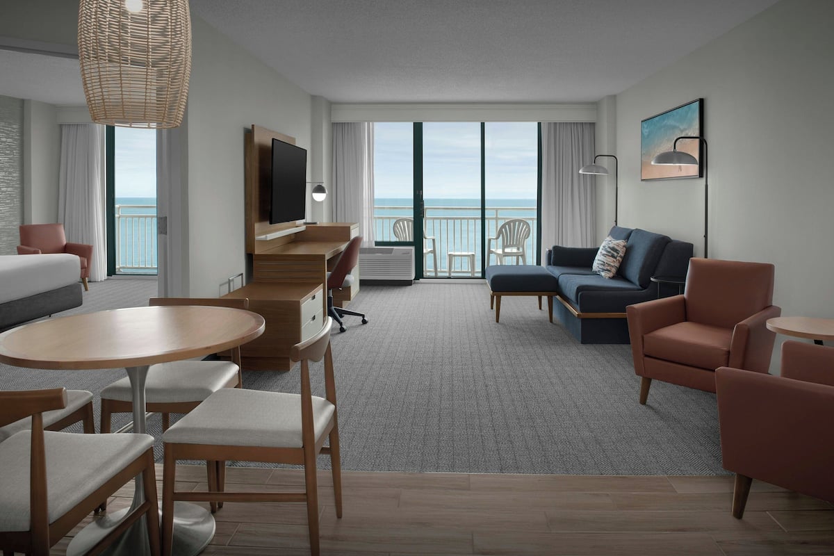 A modern hotel room with a balcony, a sofa, a dining area, and a TV.