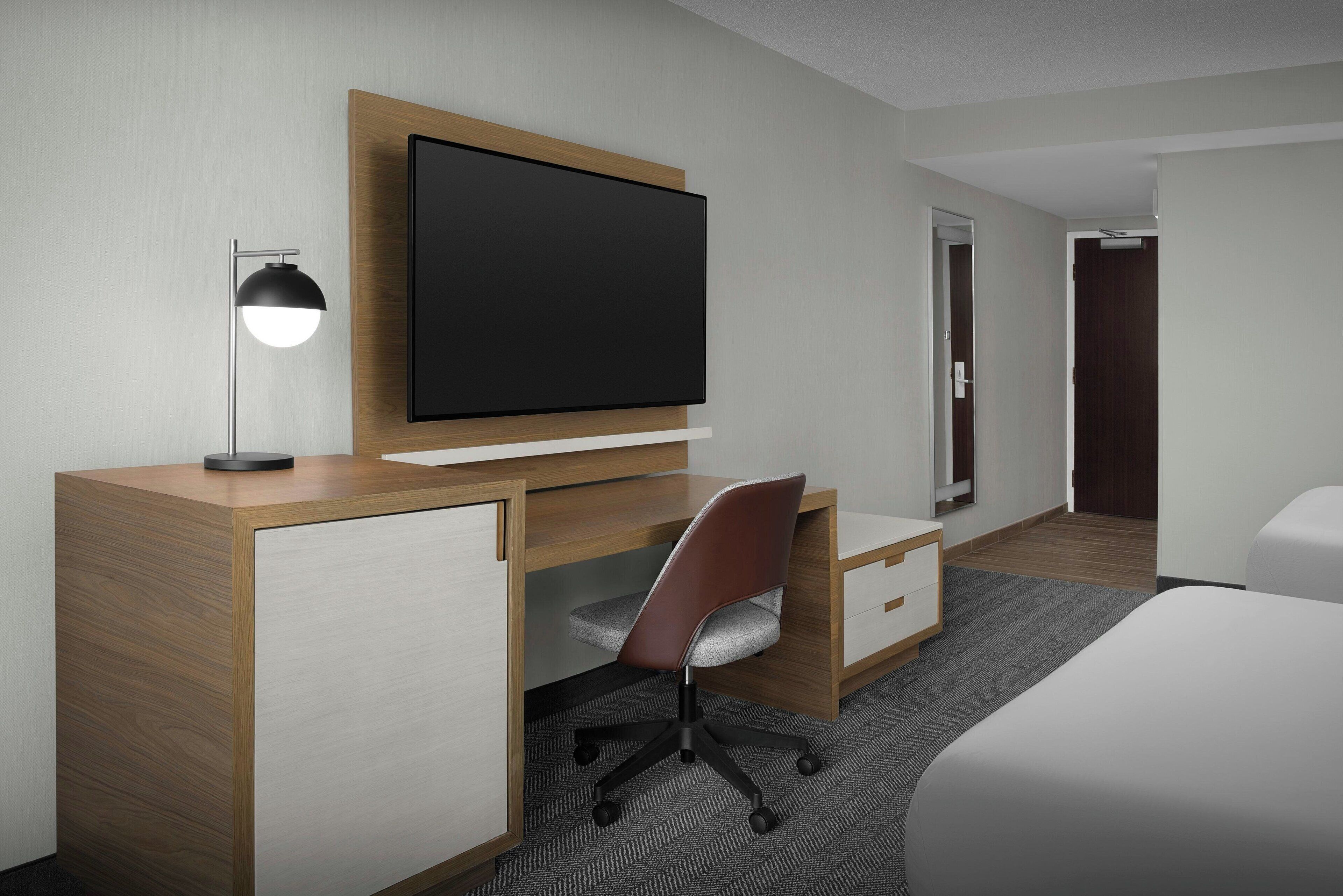 A modern hotel room with a wooden desk, a flat-screen TV, a desk lamp, a cushioned chair, a bed, and a carpeted floor.