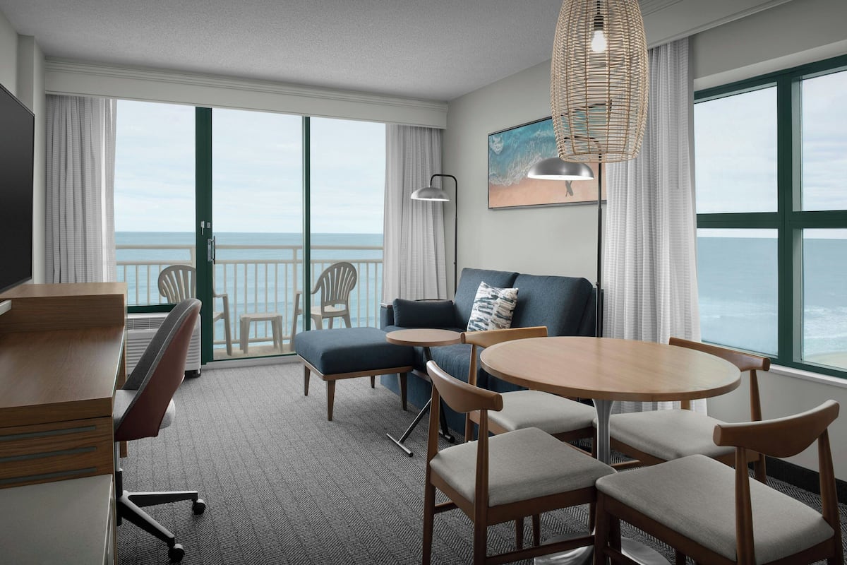 A hotel room with a balcony, a sofa, a dining table, and a TV.