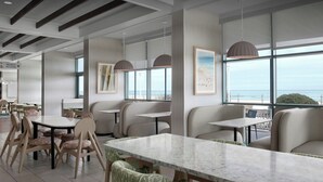 Un comedor moderno con mesas redondas y sillas acolchadas, ventanas amplias con vista al mar y un letrero que indica acceso a la playa.