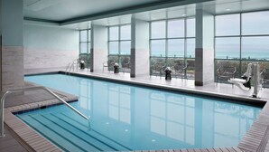 Una piscina interior con vistas despejadas al océano a través de amplios ventanales, suelo de baldosas y una escalera metálica.