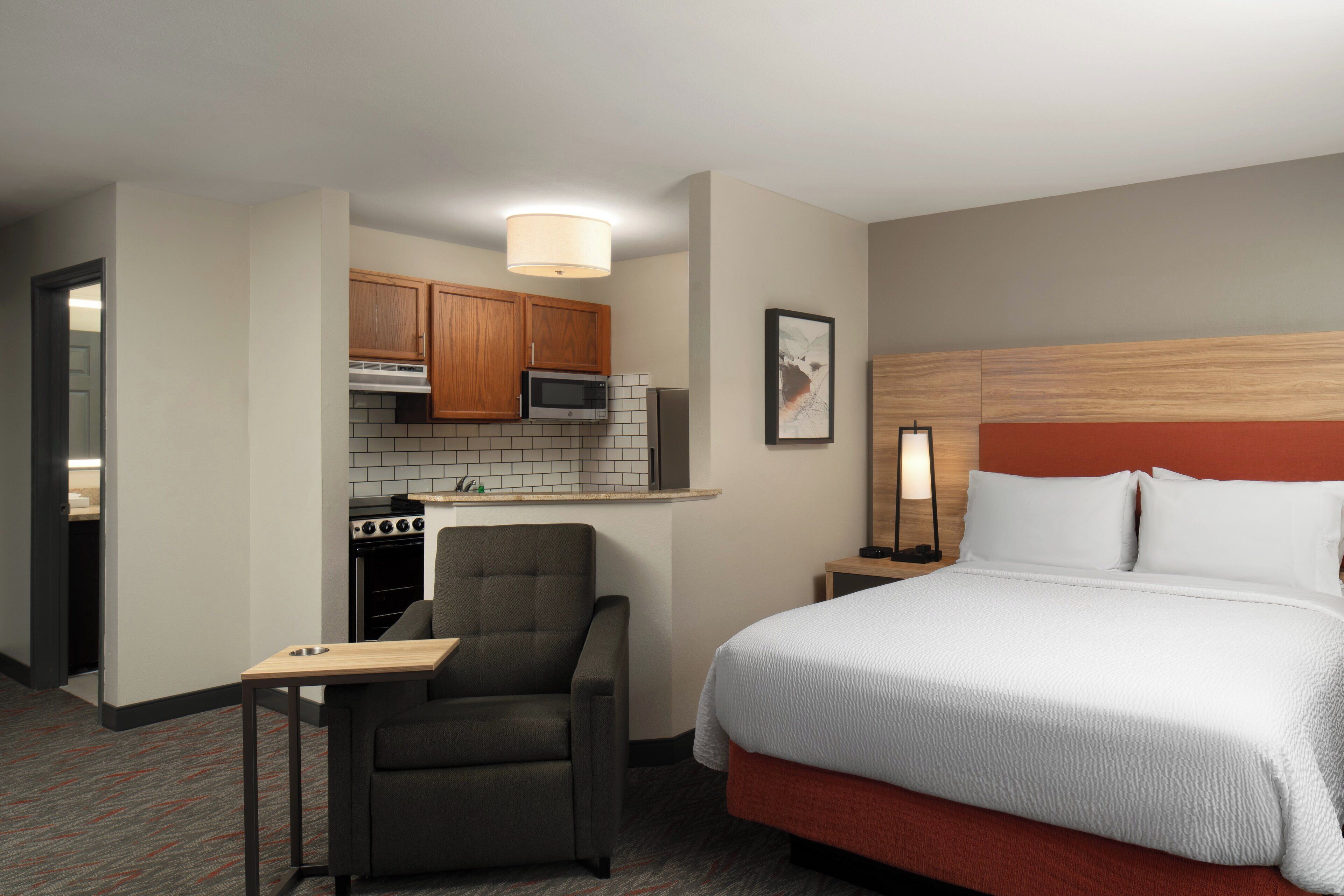 A hotel room with a bed, a chair, a small table, a microwave, a stove, and a nightstand with a lamp.
