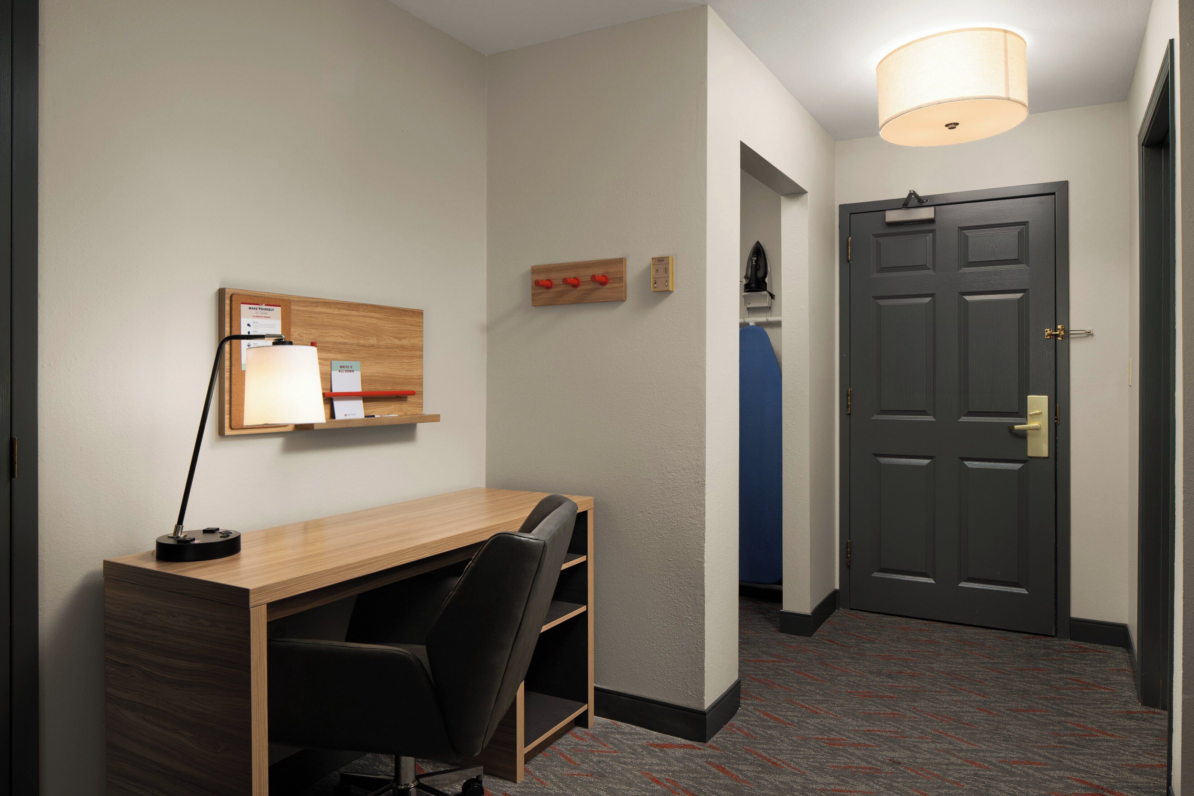 A hotel room with a wooden desk, a black office chair, a wall-mounted lamp, a bulletin board, and a dark door.