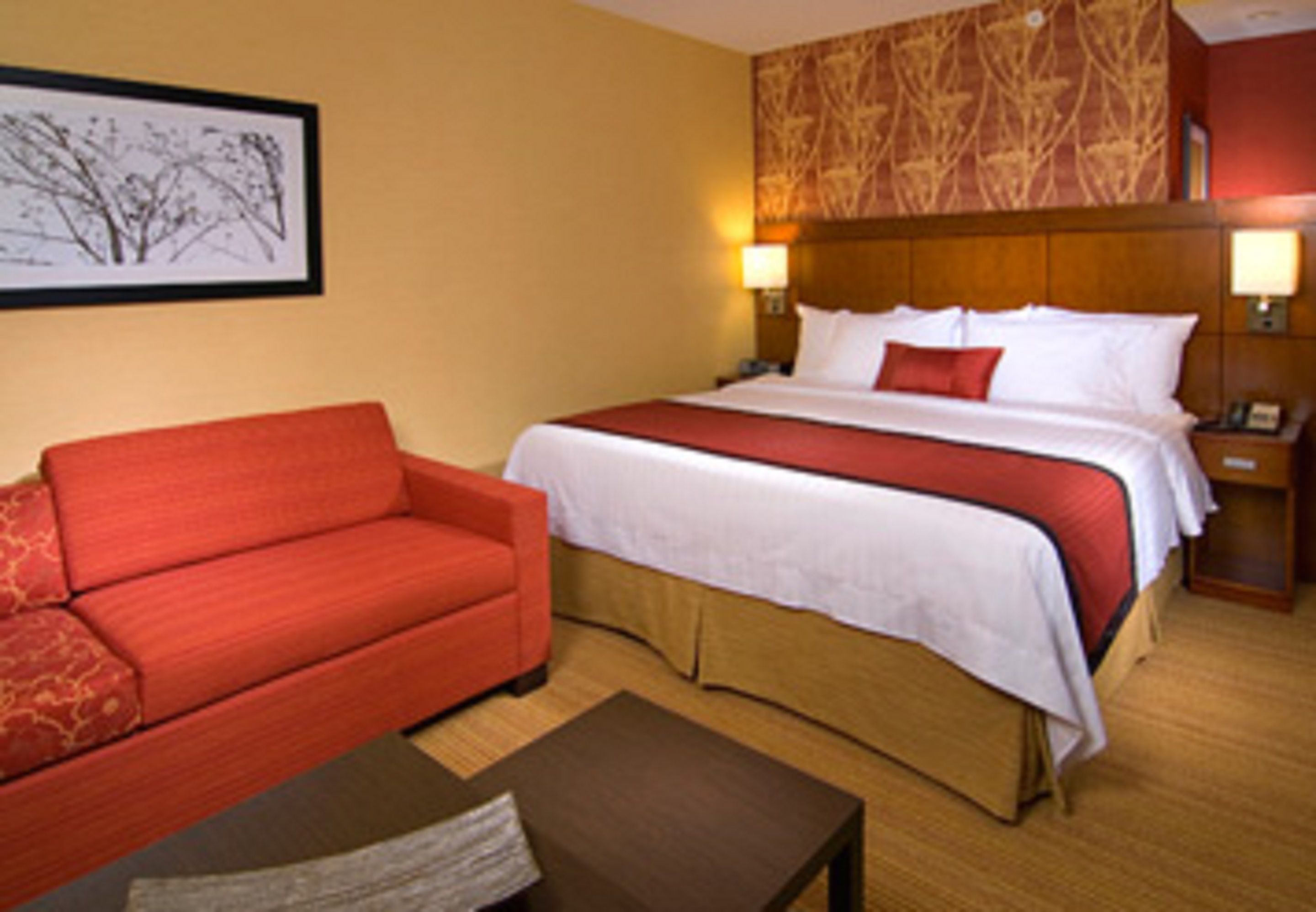 A hotel room with a bed, a red sofa, a wooden nightstand, and a framed picture on the wall.