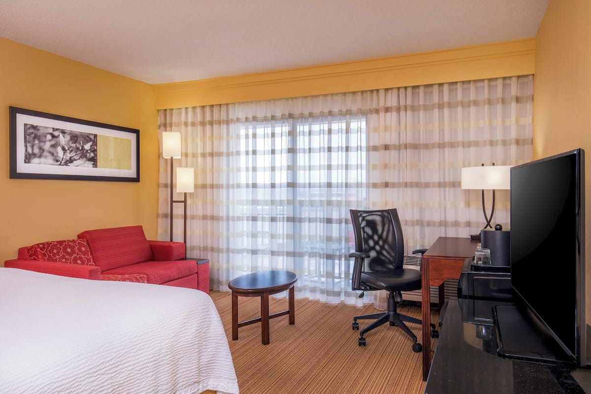 Une chambre d’hôtel avec un lit, un canapé rouge, un bureau avec un ordinateur, une télévision et une fenêtre avec des rideaux.