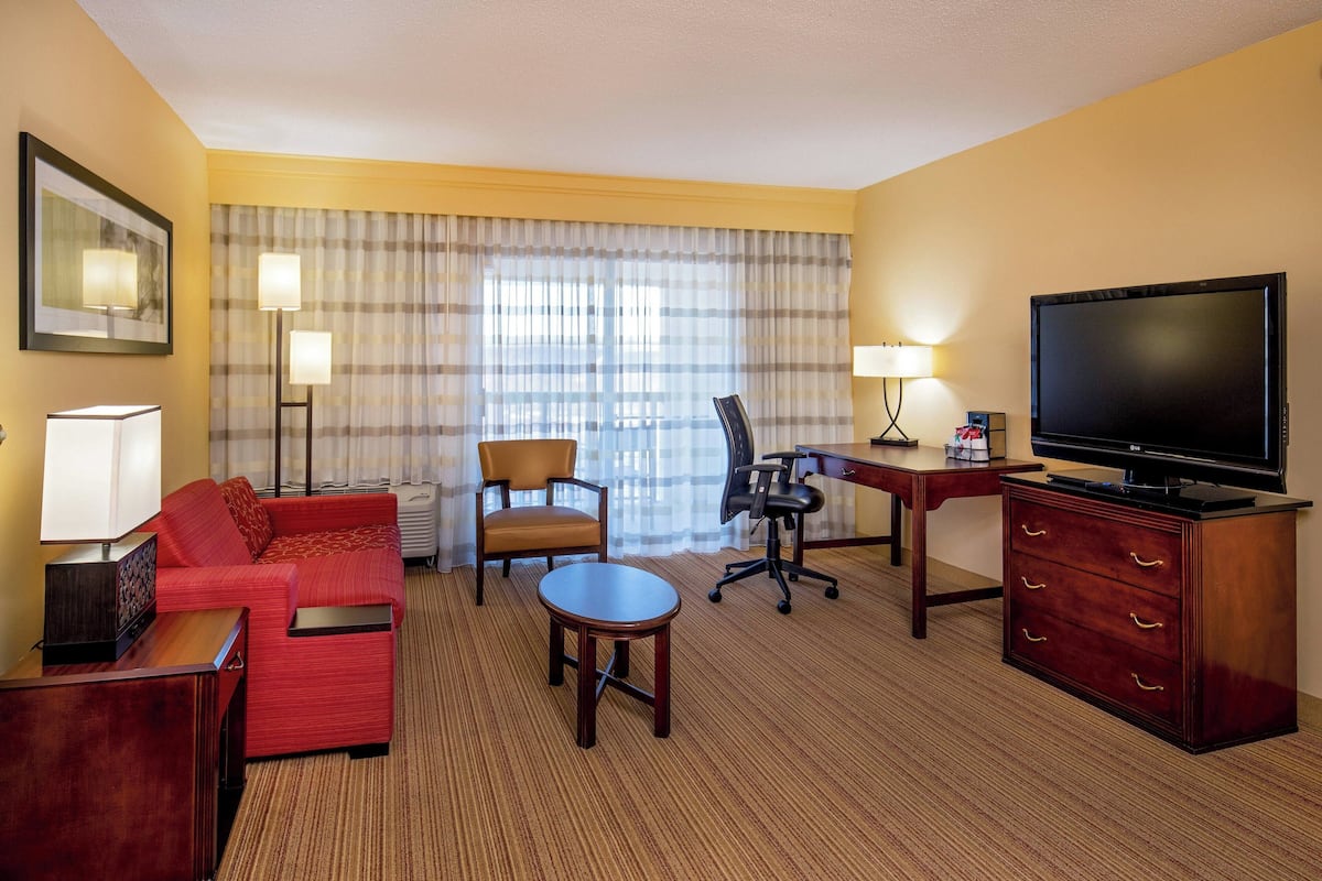 Une chambre d’hôtel avec un canapé rouge, une petite table ronde, un bureau avec une chaise, une télévision et une grande fenêtre avec des rideaux.