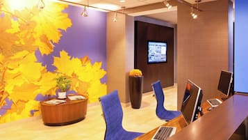 Un área de recepción con un escritorio de madera, sillas azules, una planta en maceta, una pantalla grande y una pared decorada con hojas otoñales.