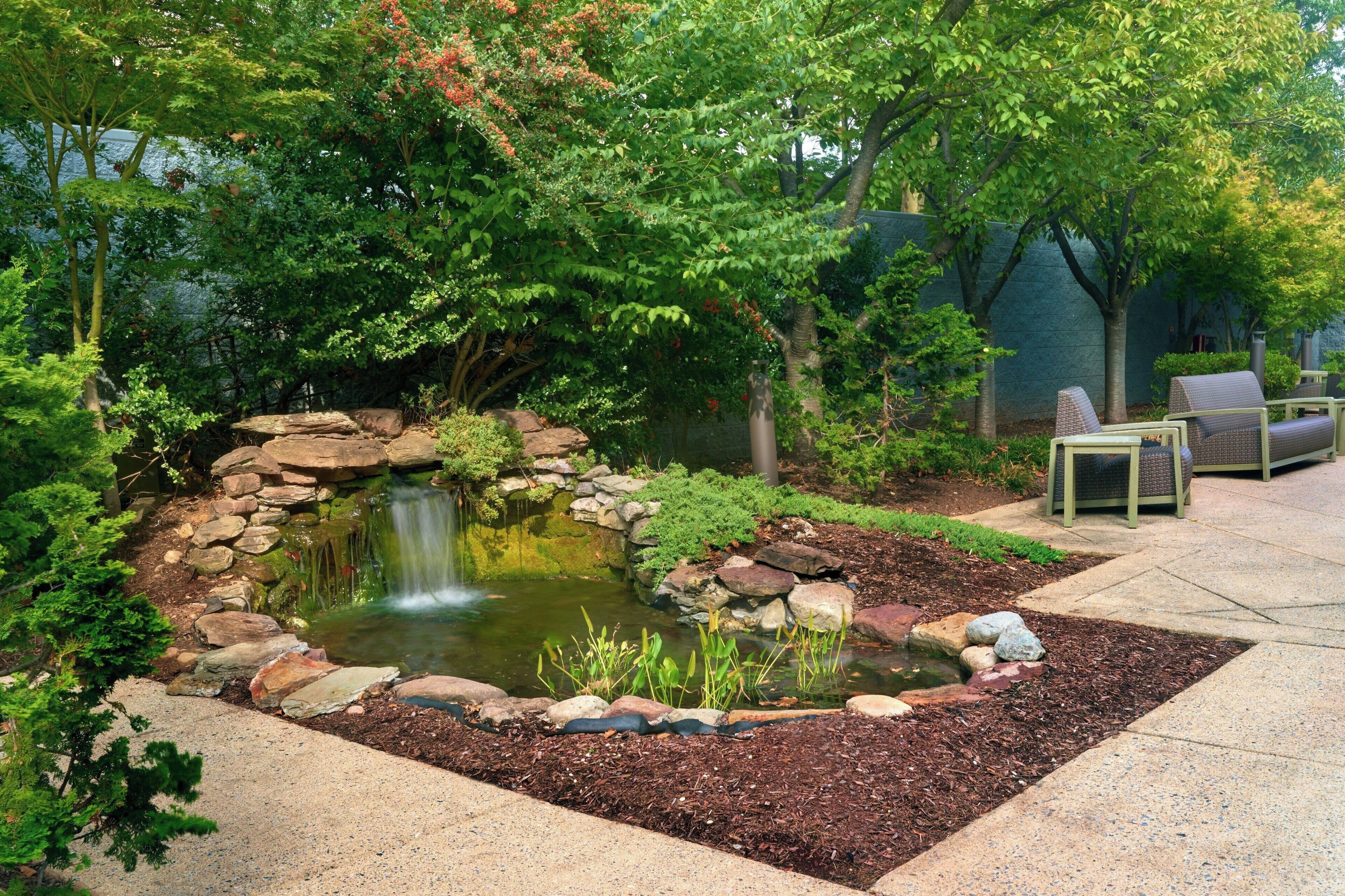 A garden with a pond, rocks, and a small waterfall. There are trees and a patio with chairs.