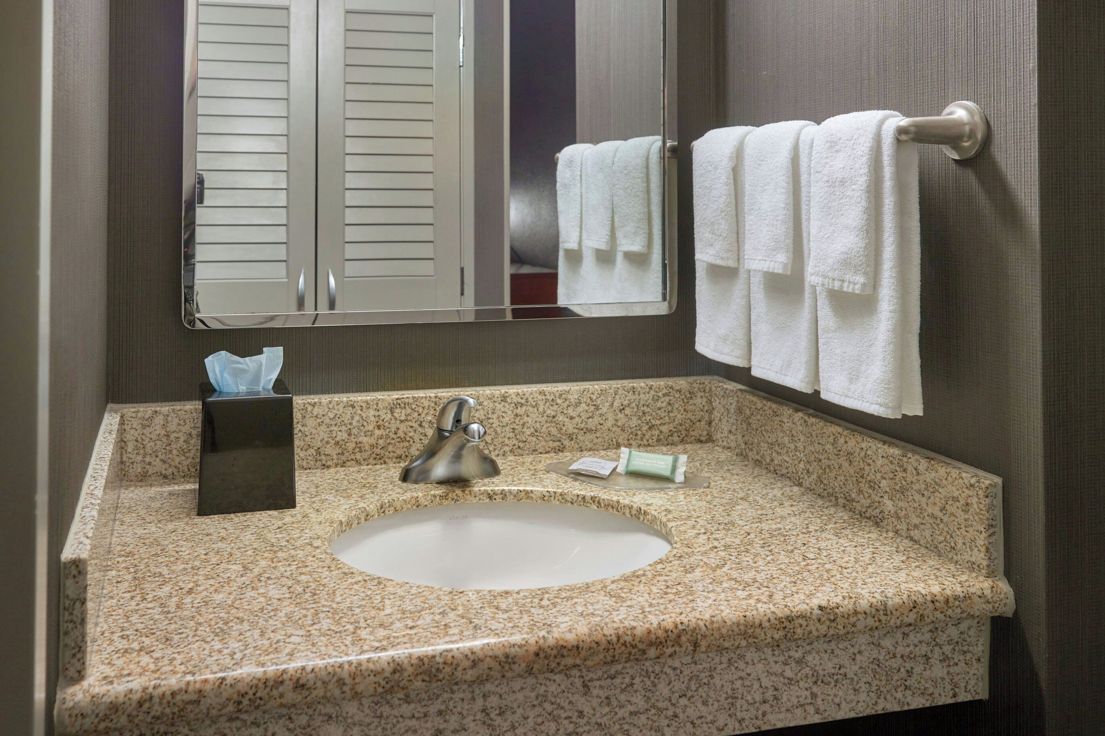 A bathroom with a granite countertop sink, a mirror, a towel rack with white towels, a soap dispenser, and a tissue box.