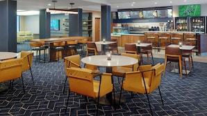 Ein modernes Restaurant mit runden Tischen und gelben Stühlen, einem Barbereich und einem Fernseher, der eine Speisekarte zeigt.