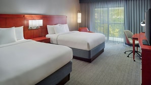 Ein Hotelzimmer mit zwei Betten, einem Schreibtisch, einem Sessel, einem Fernseher und einem Vorhangfenster.