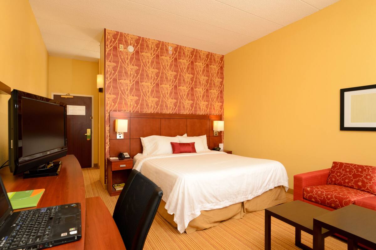 Une chambre d’hôtel avec un lit, un bureau avec un ordinateur portable, une télévision, un canapé rouge et une tête de lit en bois.