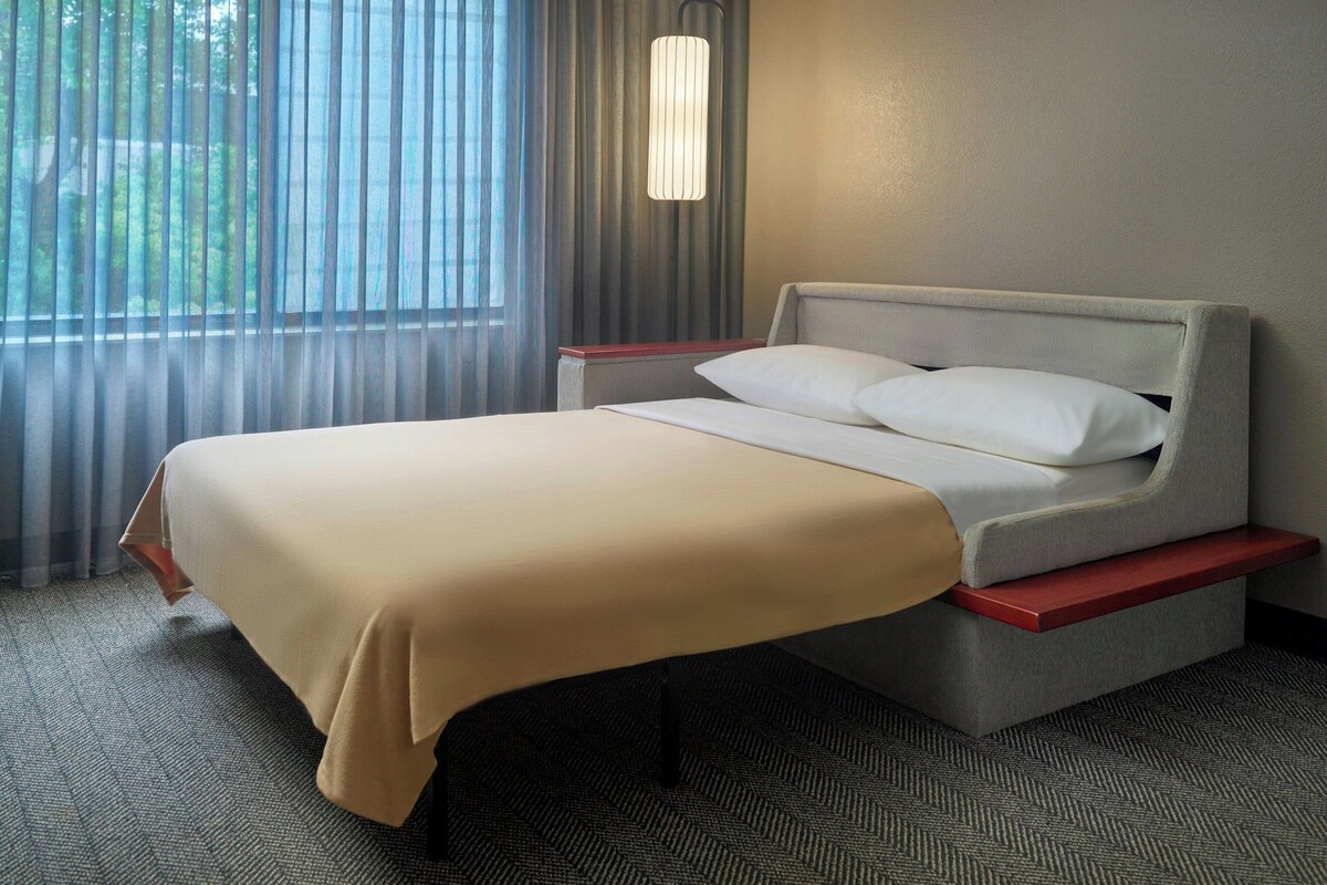 Habitación de hotel con cama, mesita de noche, lámpara y vistas al exterior por la ventana.