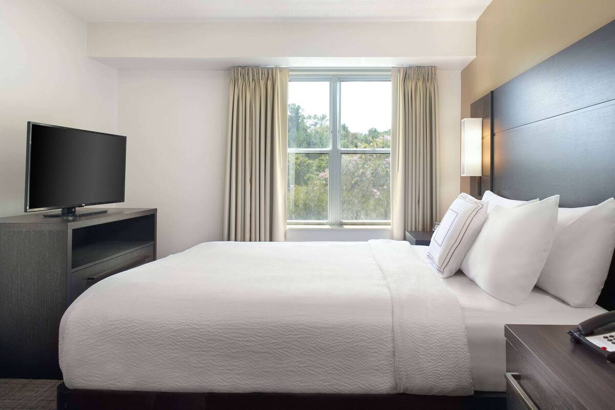 A hotel room with a large bed, a TV on a stand, a bedside table, and a window with curtains.