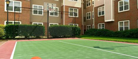 Una cancha de baloncesto al aire libre con aro y balón, rodeada por un edificio de ladrillo con varias ventanas y un seto.