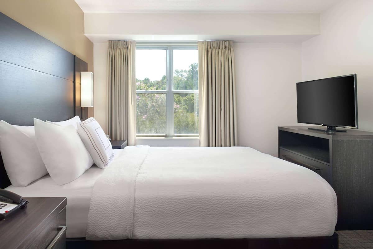 A hotel room with a large bed, a TV on a stand, a bedside table with a phone, and a window with curtains.