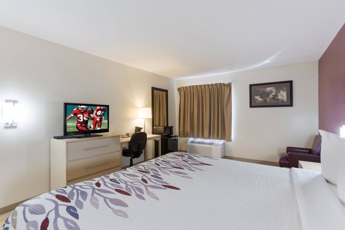 Quarto de hotel com cama, escrivaninha com computador, televisão, cadeira e janela com cortinas.