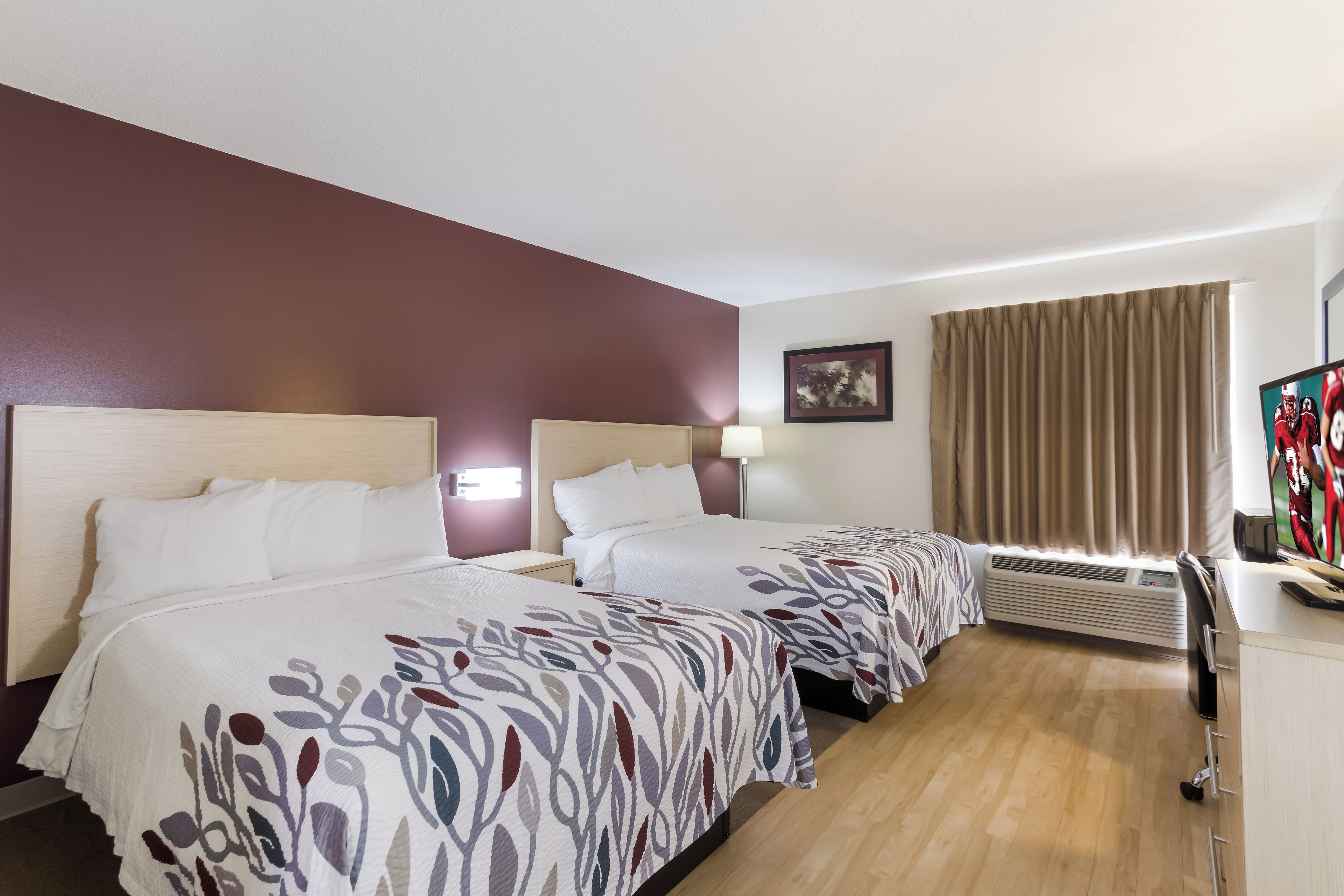 A hotel room with two beds, a desk with a computer, a window with curtains, and a wall-mounted air conditioner.