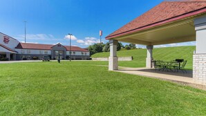 Un bâtiment doté d’un espace extérieur couvert avec des sièges, d’un parking et d’un terrain herbeux.