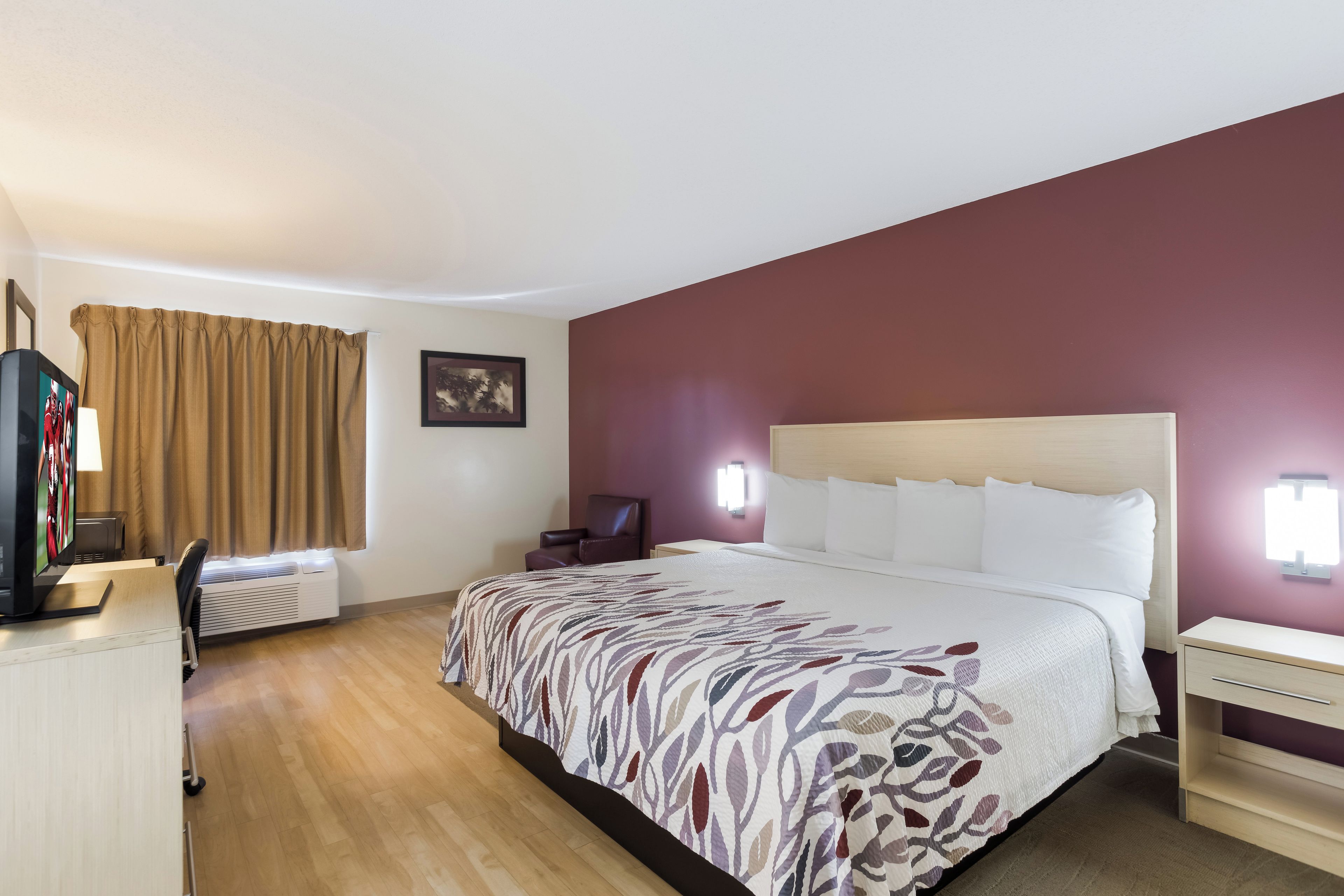 Una habitación de hotel con cama, escritorio, silla, televisor y ventana con cortinas.