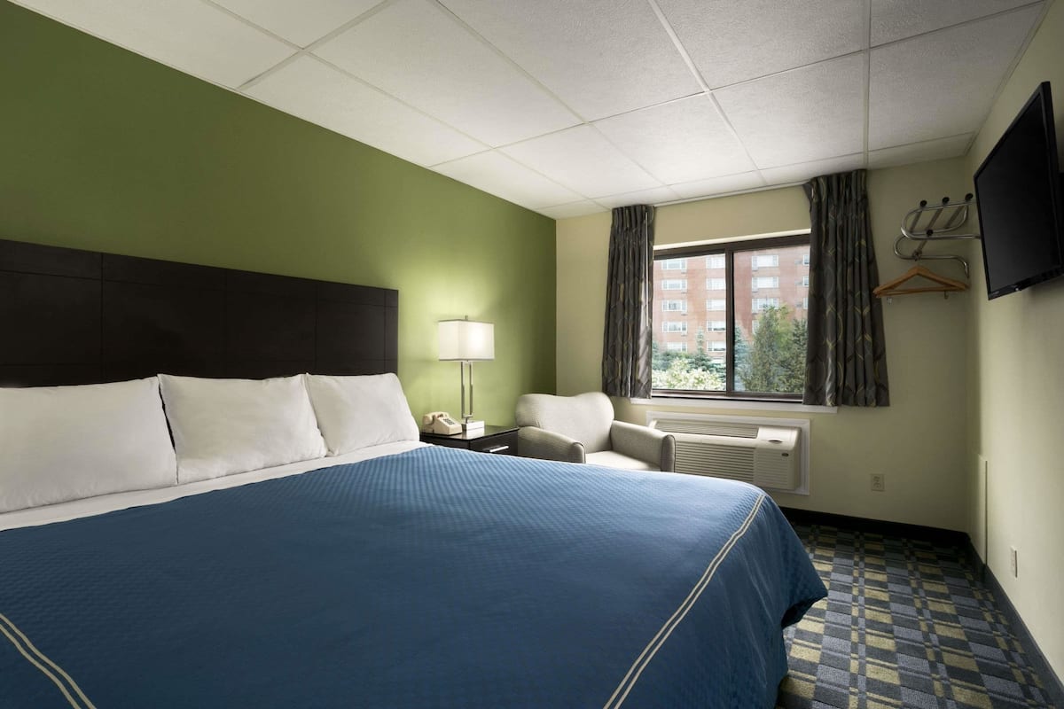 A hotel room with a large bed, a chair, a desk with a lamp, a TV, and a window with curtains.
