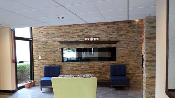 A room with a stone wall, a flat-screen TV mounted above a fireplace, two blue armchairs, and a yellow table.