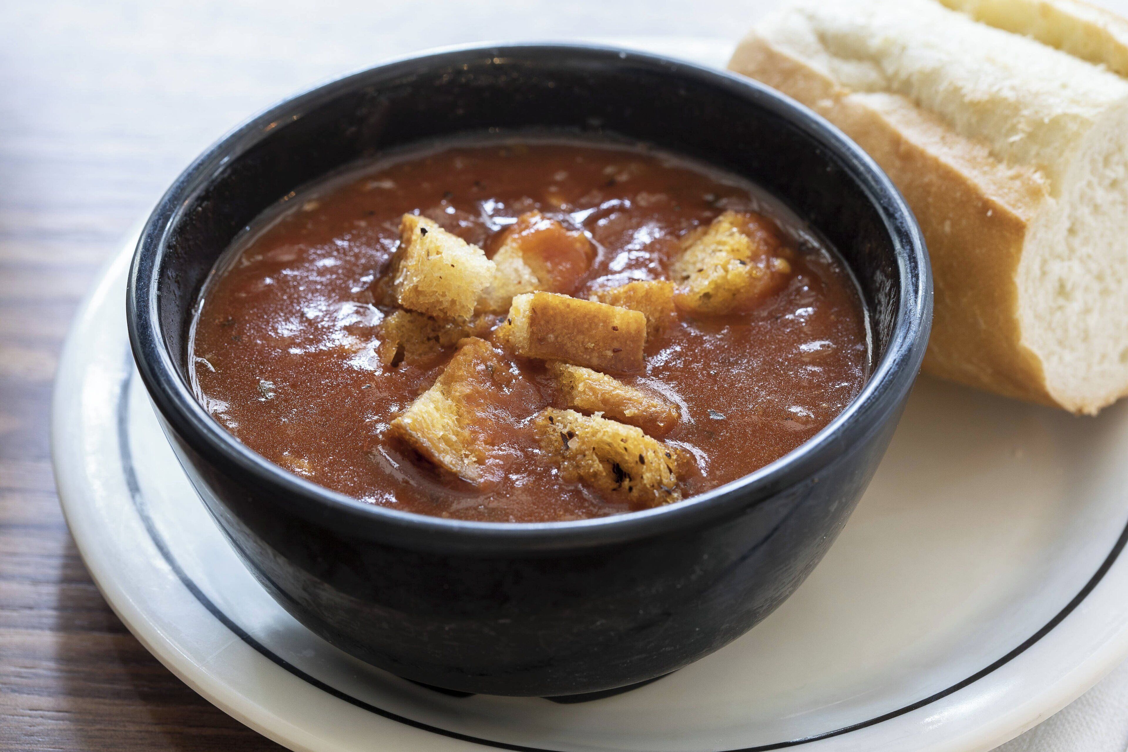 A bowl of soup with croutons and a slice of bread on a plate.