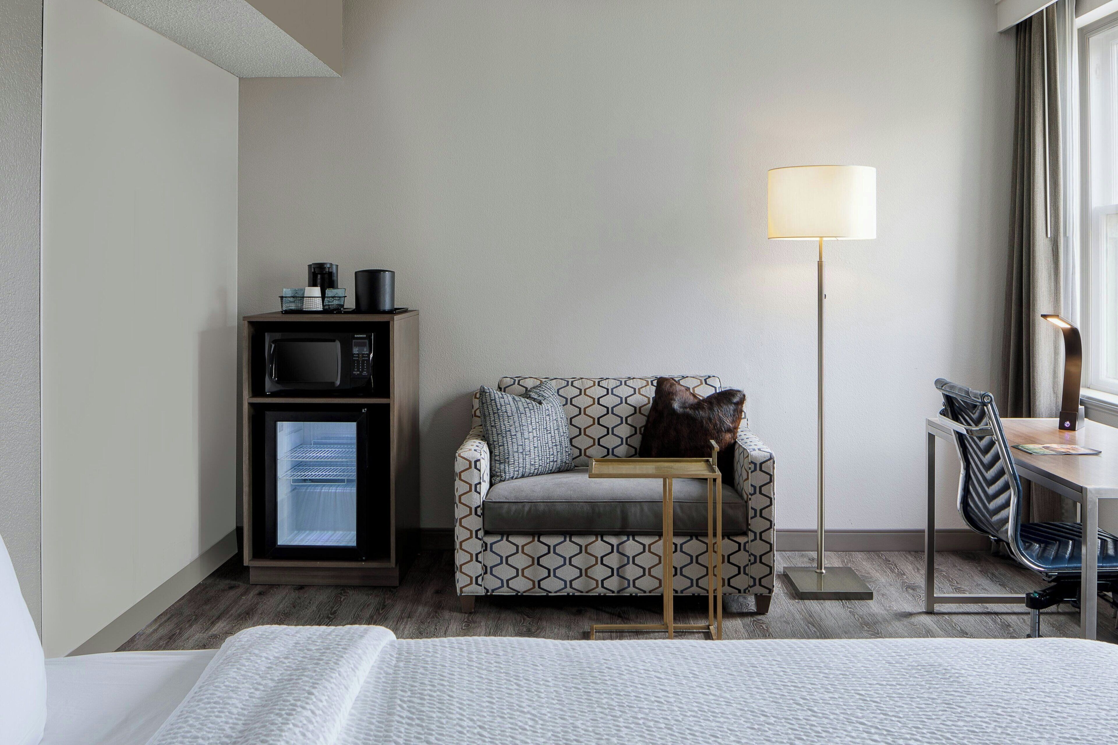 A hotel room with a bed, a sofa, a desk, a lamp, a coffee machine, and a mini-fridge.