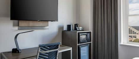 Habitación de hotel con televisión de pantalla plana montada en la pared, un escritorio con silla, microondas y refrigerador pequeño.