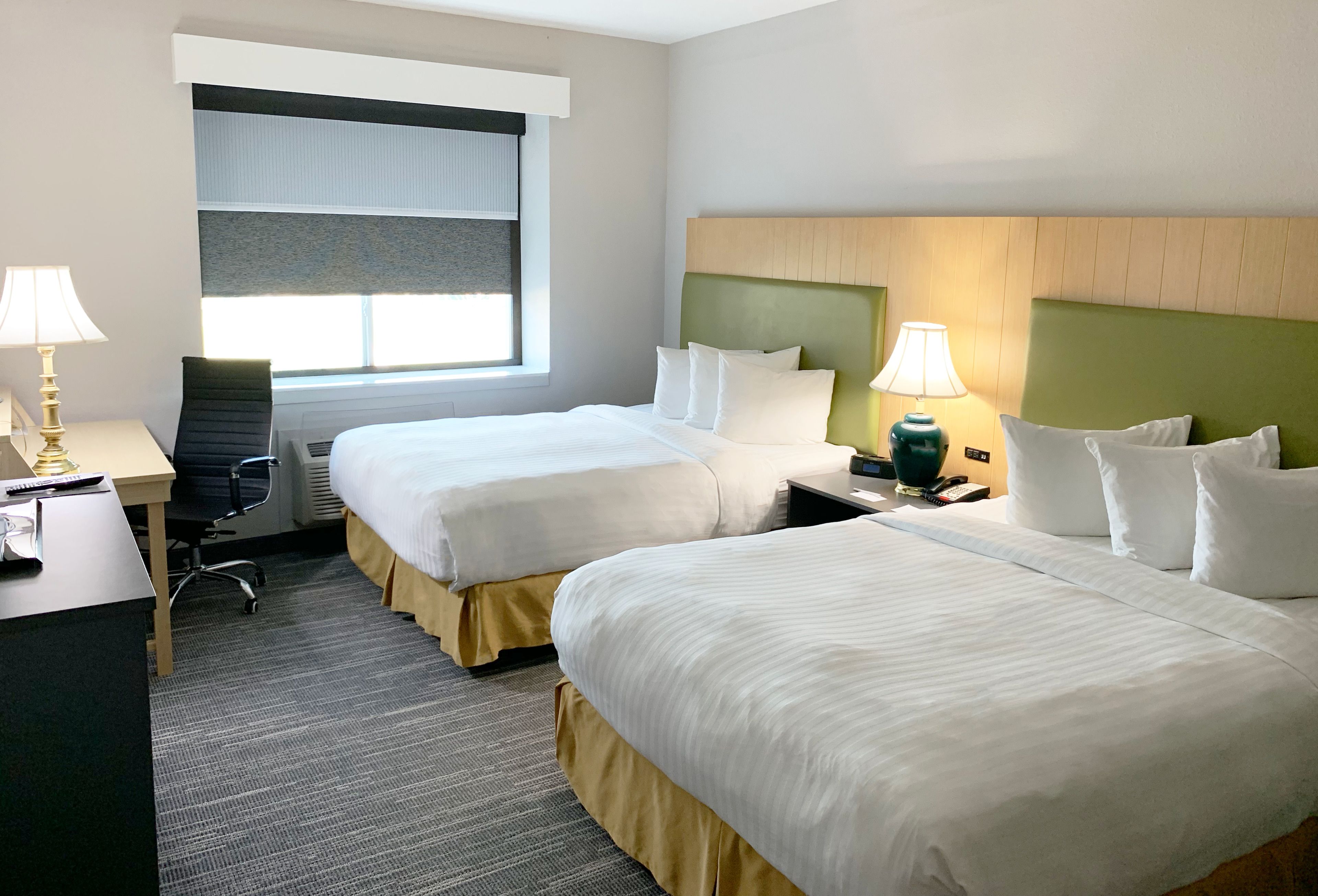 A hotel room with two beds, a desk, a chair, a lamp, and a window with blinds.