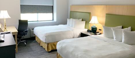 A hotel room with two beds, a desk, a chair, a lamp, and a window with blinds.