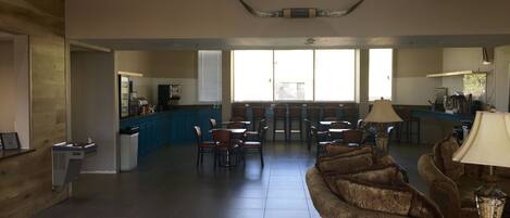 Un amplio vestíbulo de hotel con techo alto, un área de descanso central con sofás y sillones, una zona de cocina y una recepción.