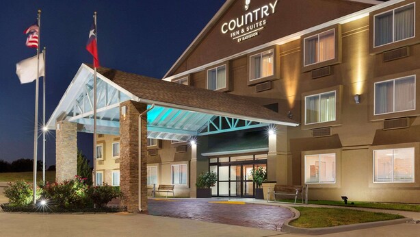 A Country Inn & Suites by Radisson building with a covered entrance, flagpoles, and a well-lit facade. - Country Inn & Suites by Radisson, Fort Worth West l-30 NAS JRB (Fort Worth)