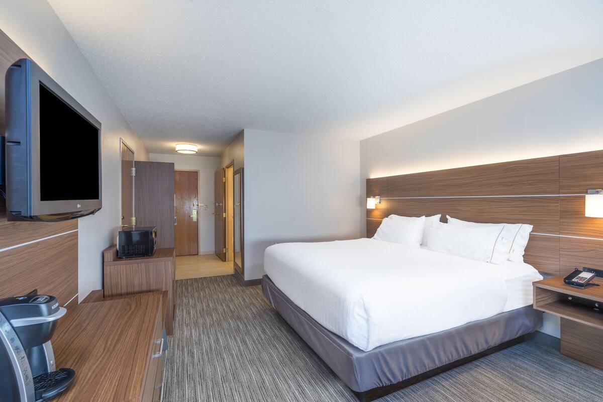 A hotel room with a large bed, a flat-screen TV mounted on the wall, a coffee machine, and a wooden headboard.