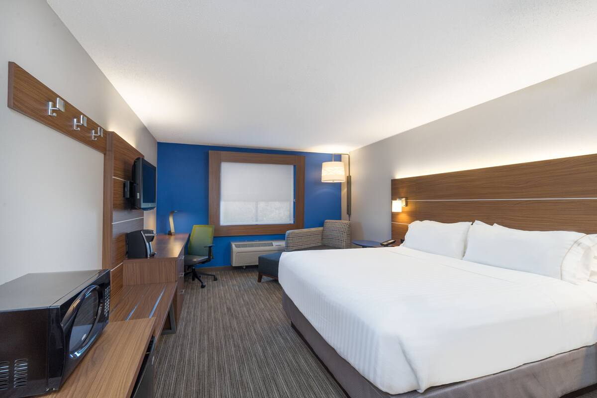 A hotel room with a large bed, a desk with a microwave, a chair, a TV, and a window.
