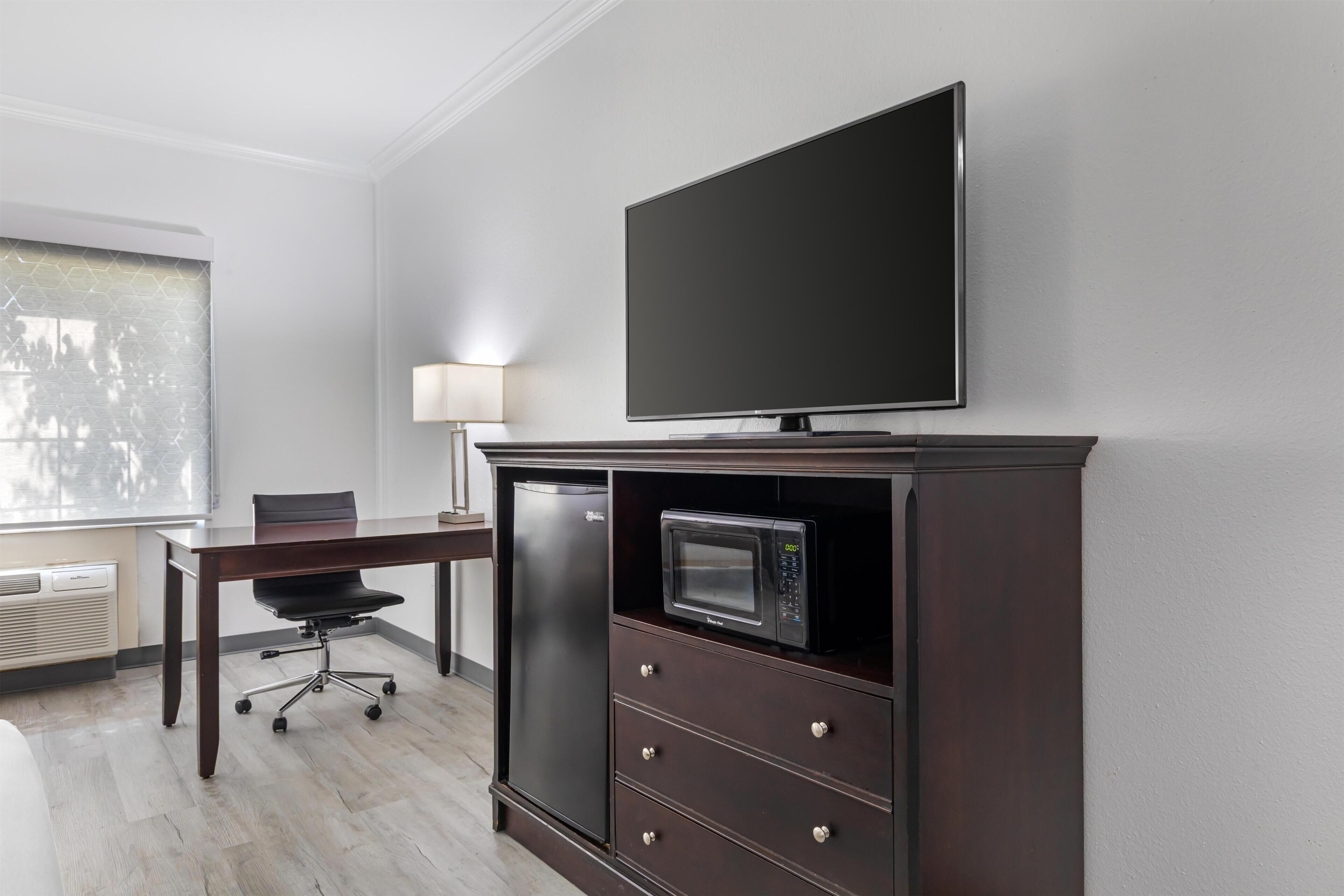Une chambre d’hôtel équipée d’un téléviseur à écran plat fixé au mur, d’un bureau avec une chaise, d’un four à micro-ondes et d’une petite armoire avec des tiroirs.
