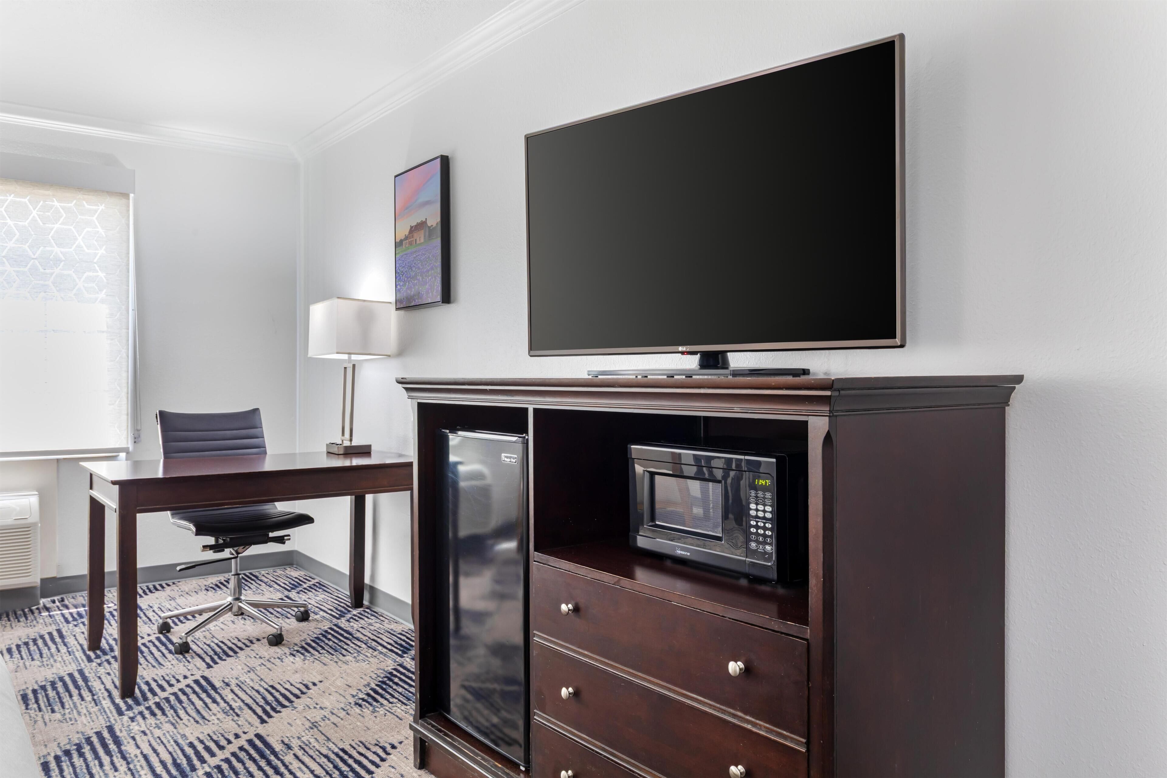 Habitación de hotel con televisión de pantalla plana, microondas, refrigerador, escritorio con silla y alfombra estampada.
